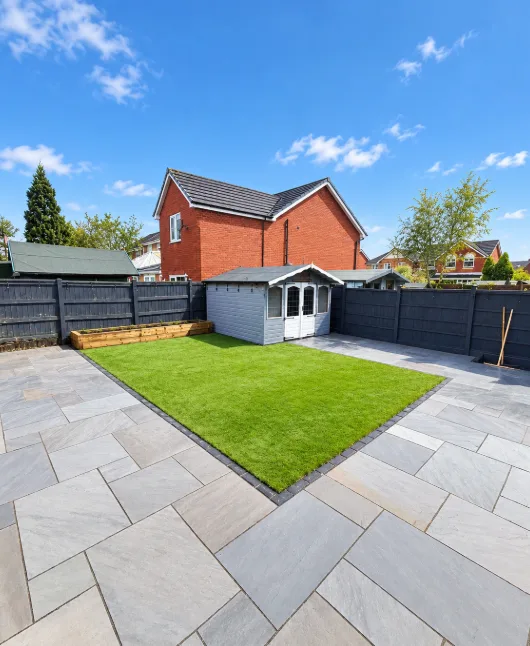 Sunny grey patio with square lawn, shed and red brick house